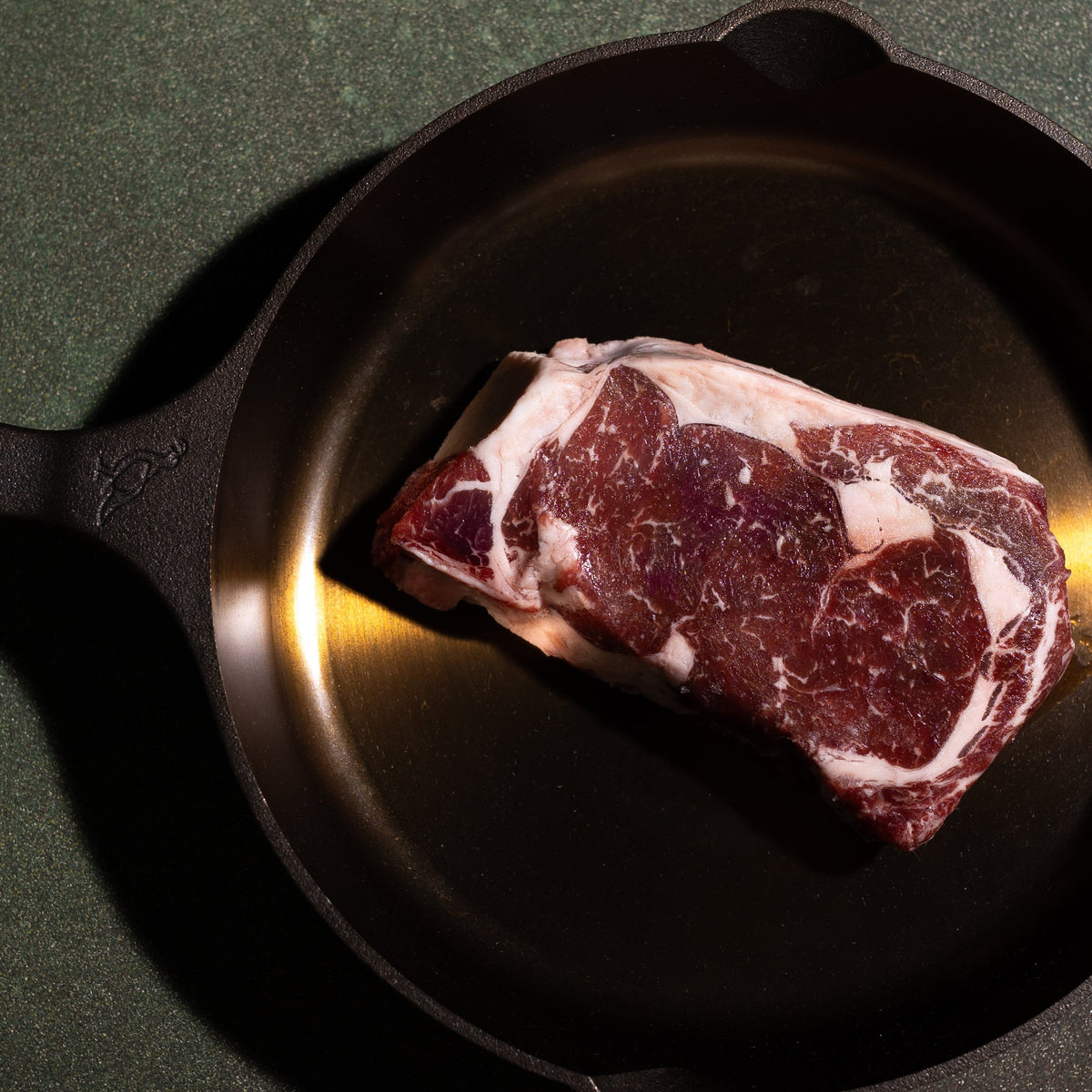 A raw cut of marbled steak placed inside a black cast iron skillet on a dark green textured surface.