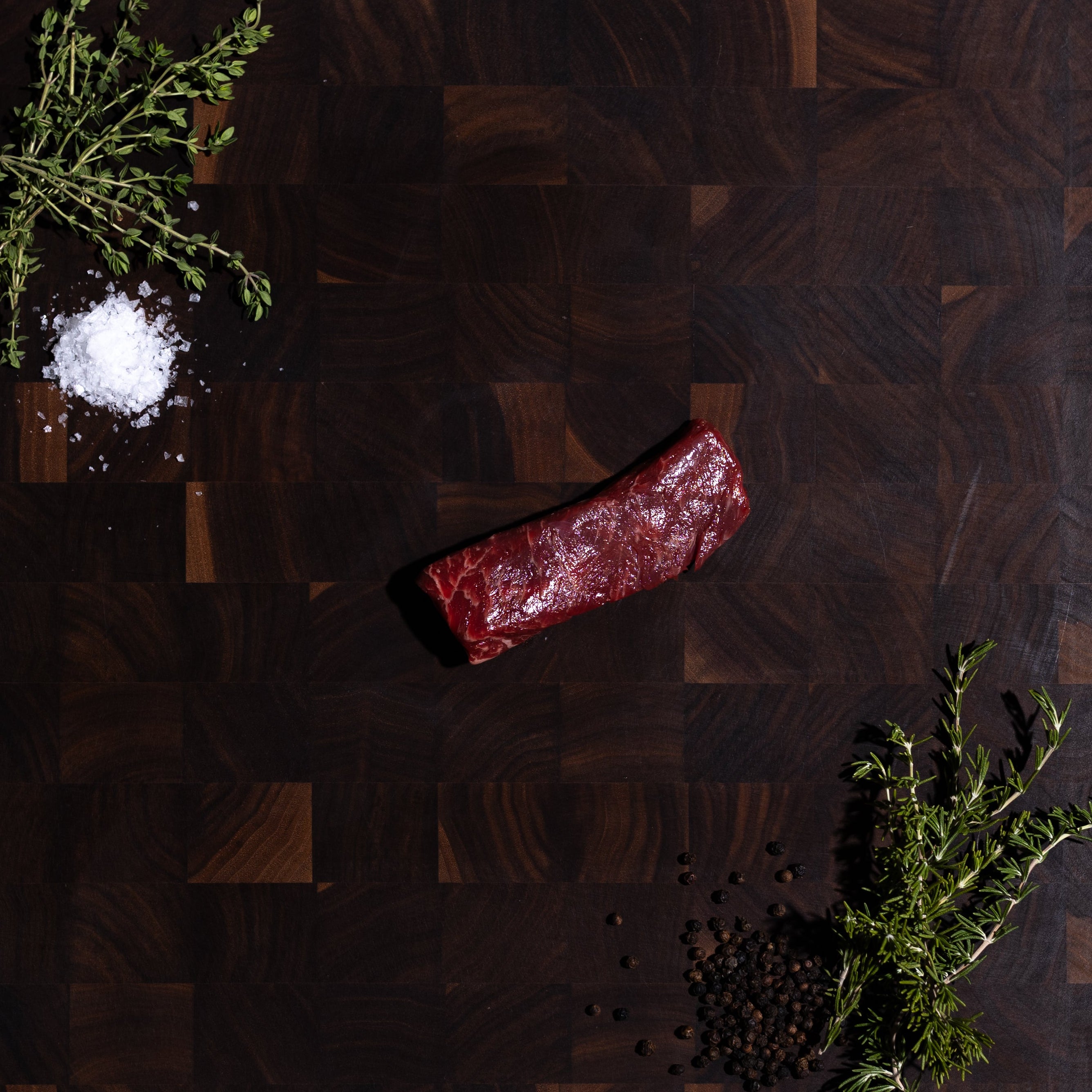 A dark brown wooden cutting board with a geometric block pattern, styled with a piece of raw meat, salt, and herbs on a dark green surface.