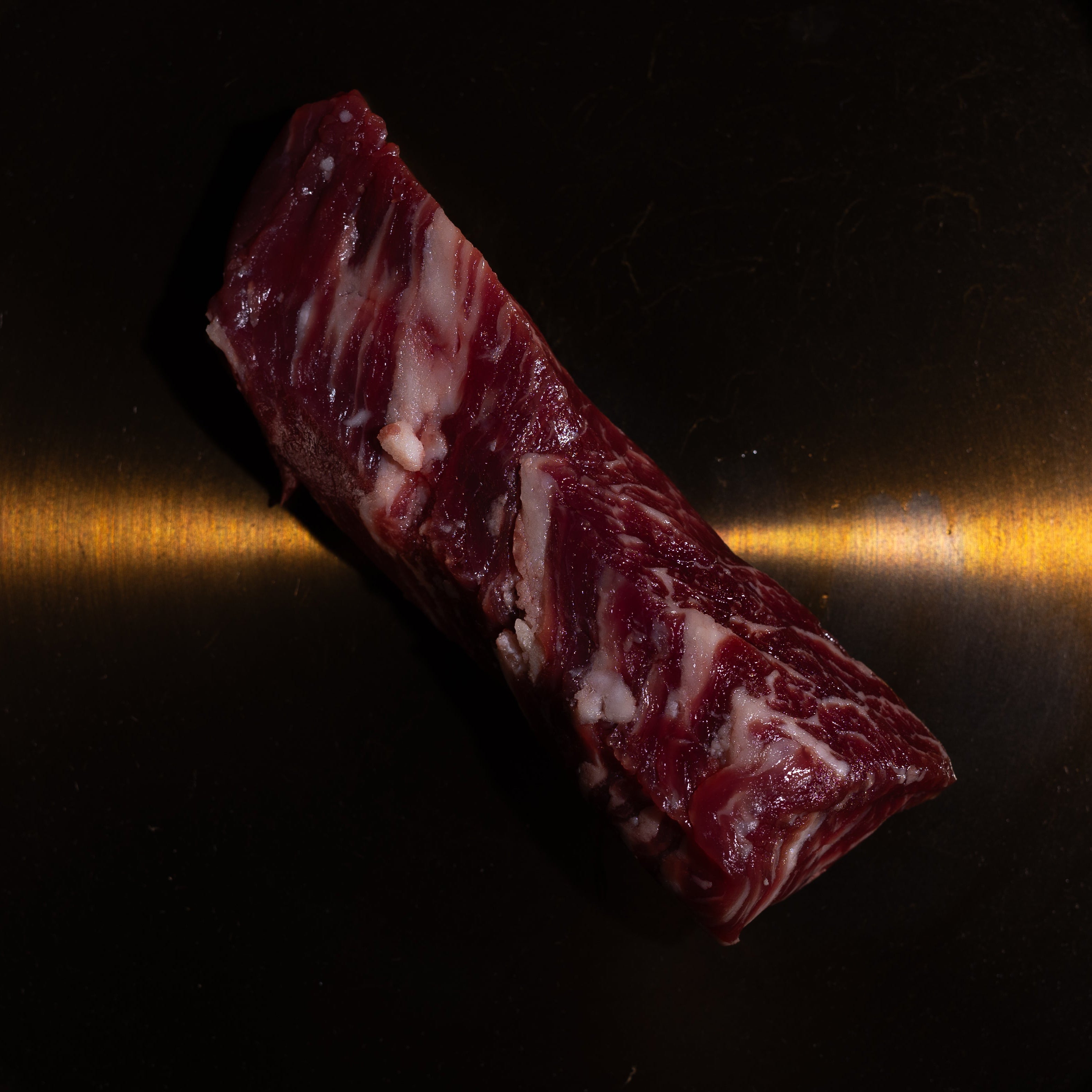 A single rectangular piece of raw marbled meat, likely beef, placed diagonally on a dark metallic pan or plate with a warm golden reflection.