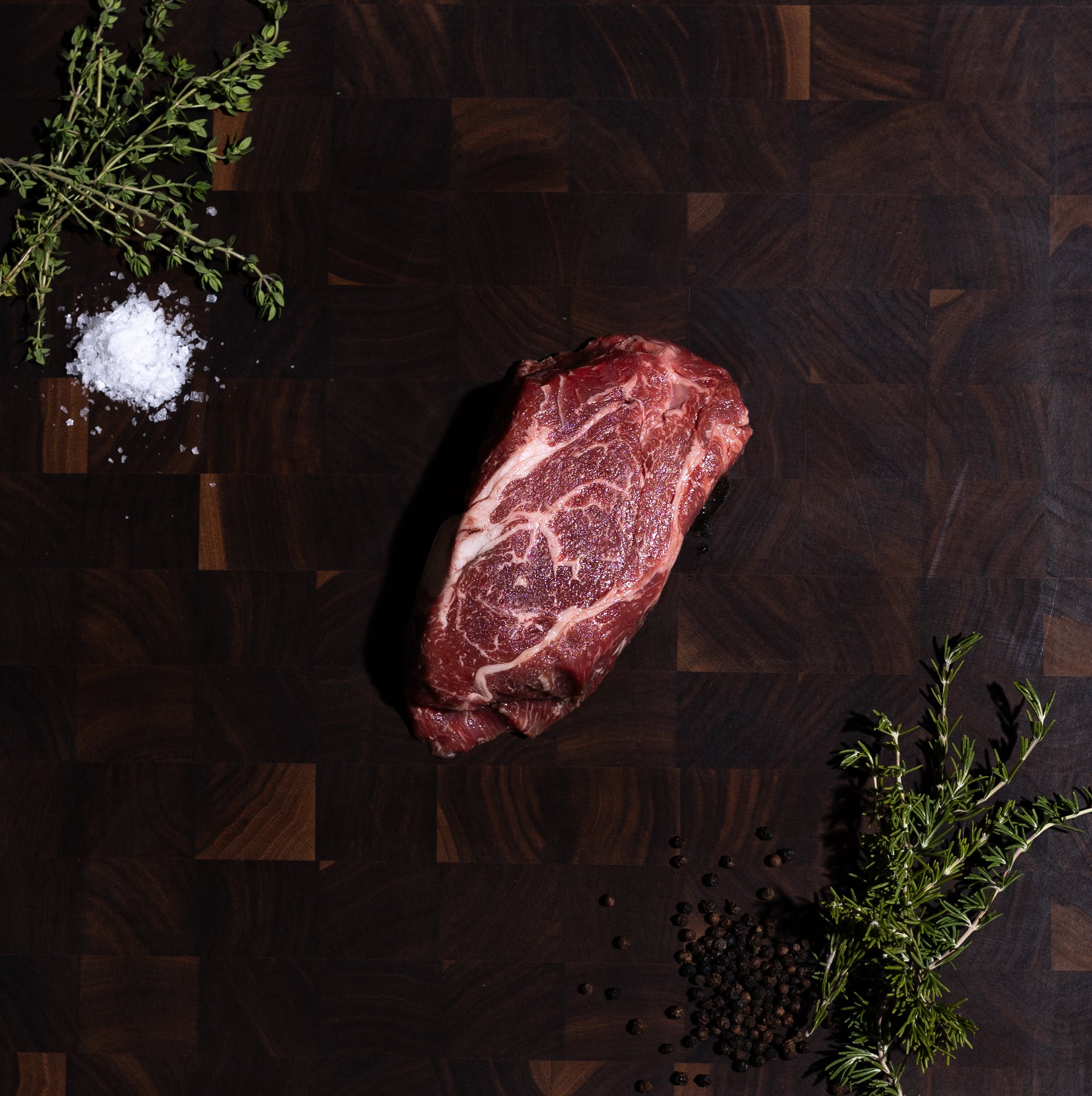 A raw marbled beef steak is placed on a dark wooden cutting board, styled with a small pile of coarse salt and green herb sprigs on a dark green surface.
