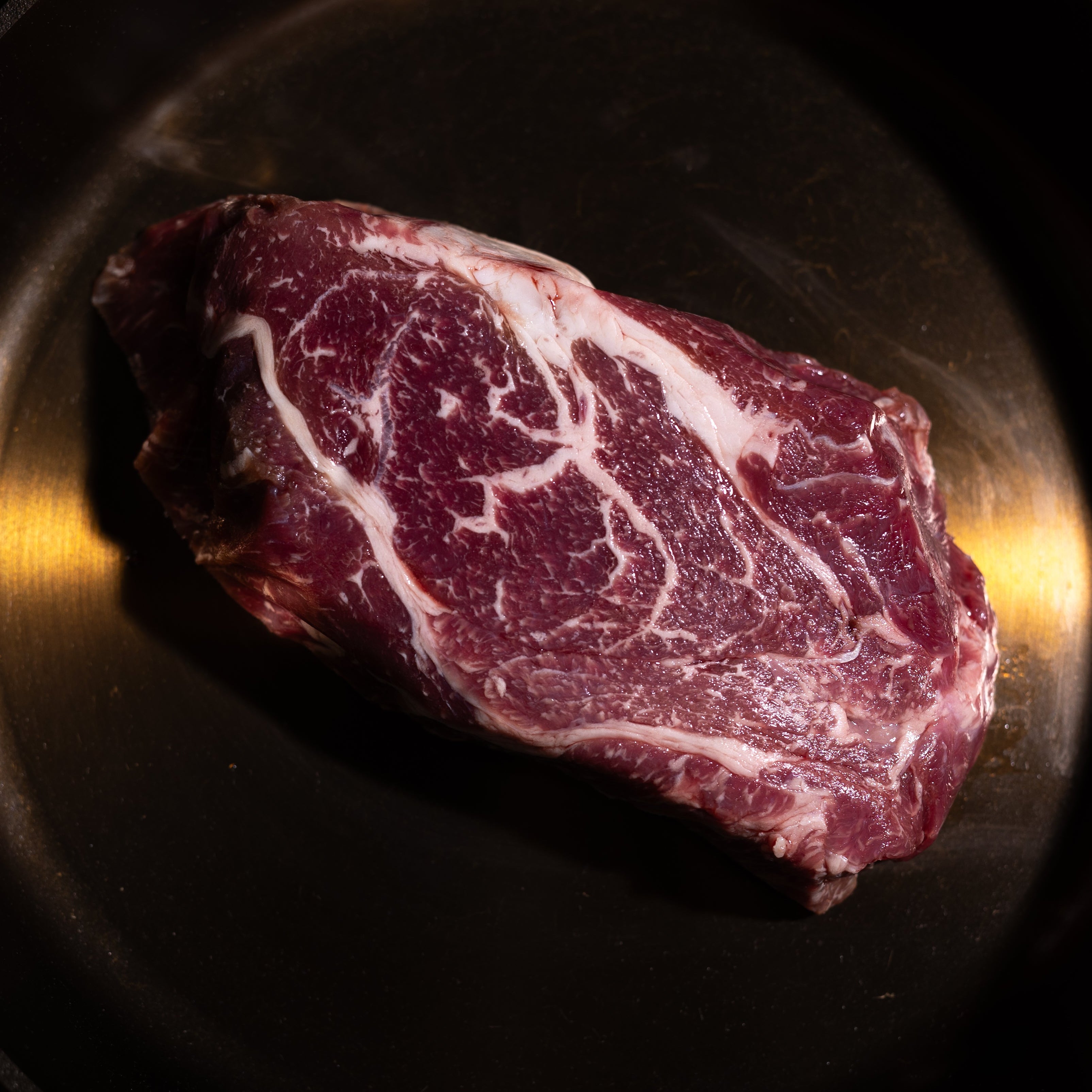A raw marbled beef steak sits inside a black cast iron skillet on a dark green textured surface.