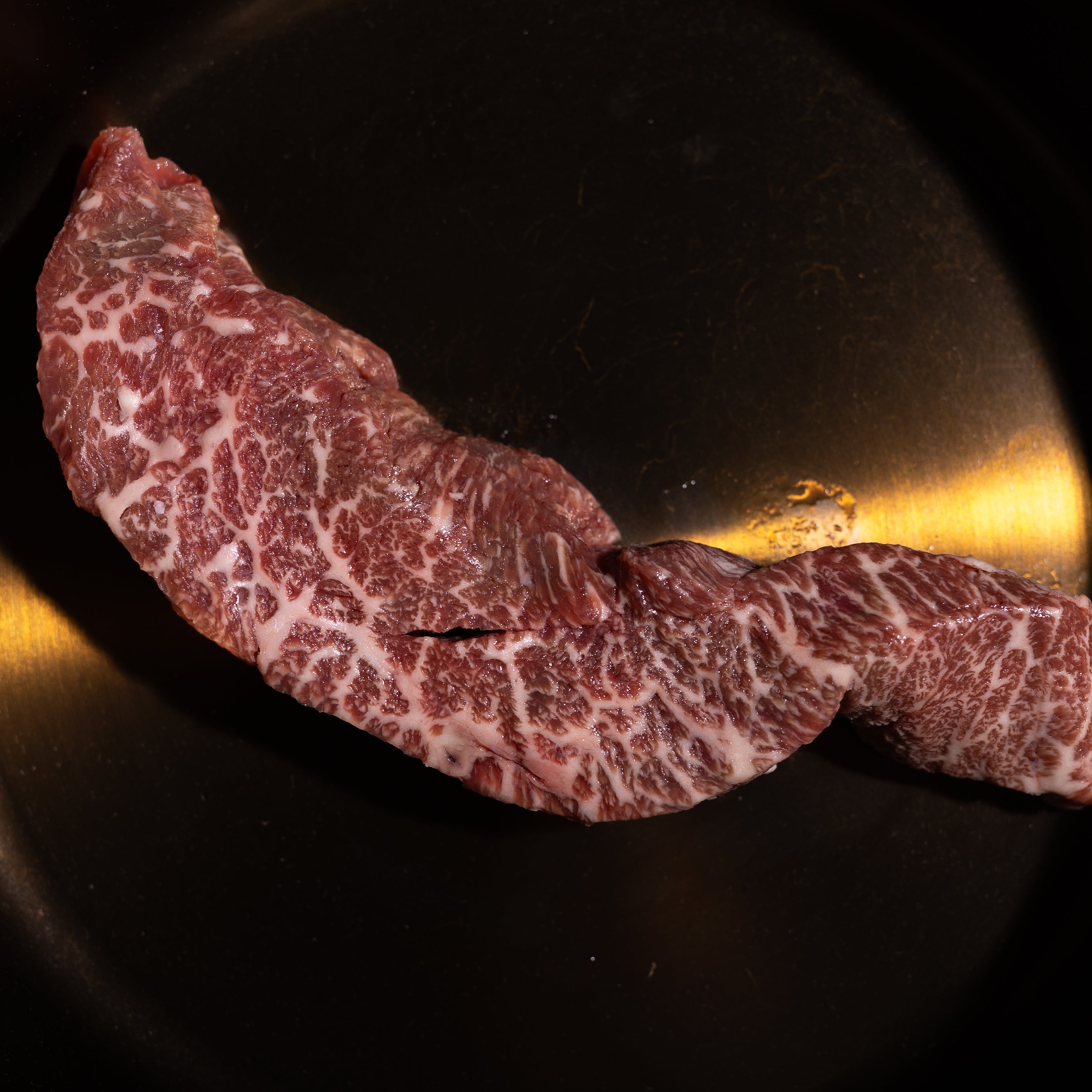 Two slices of marbled beef steak resting in a black cast iron skillet on a dark textured surface.