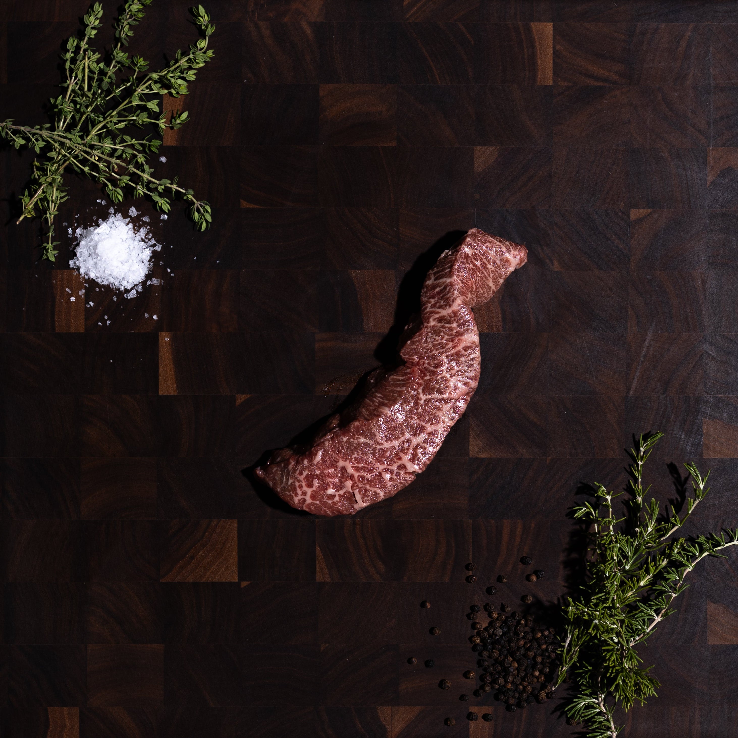 A raw cut of beef steak is arranged on a dark wooden cutting board, styled with a pinch of coarse salt, black peppercorns, and green herbs on a dark olive green background.