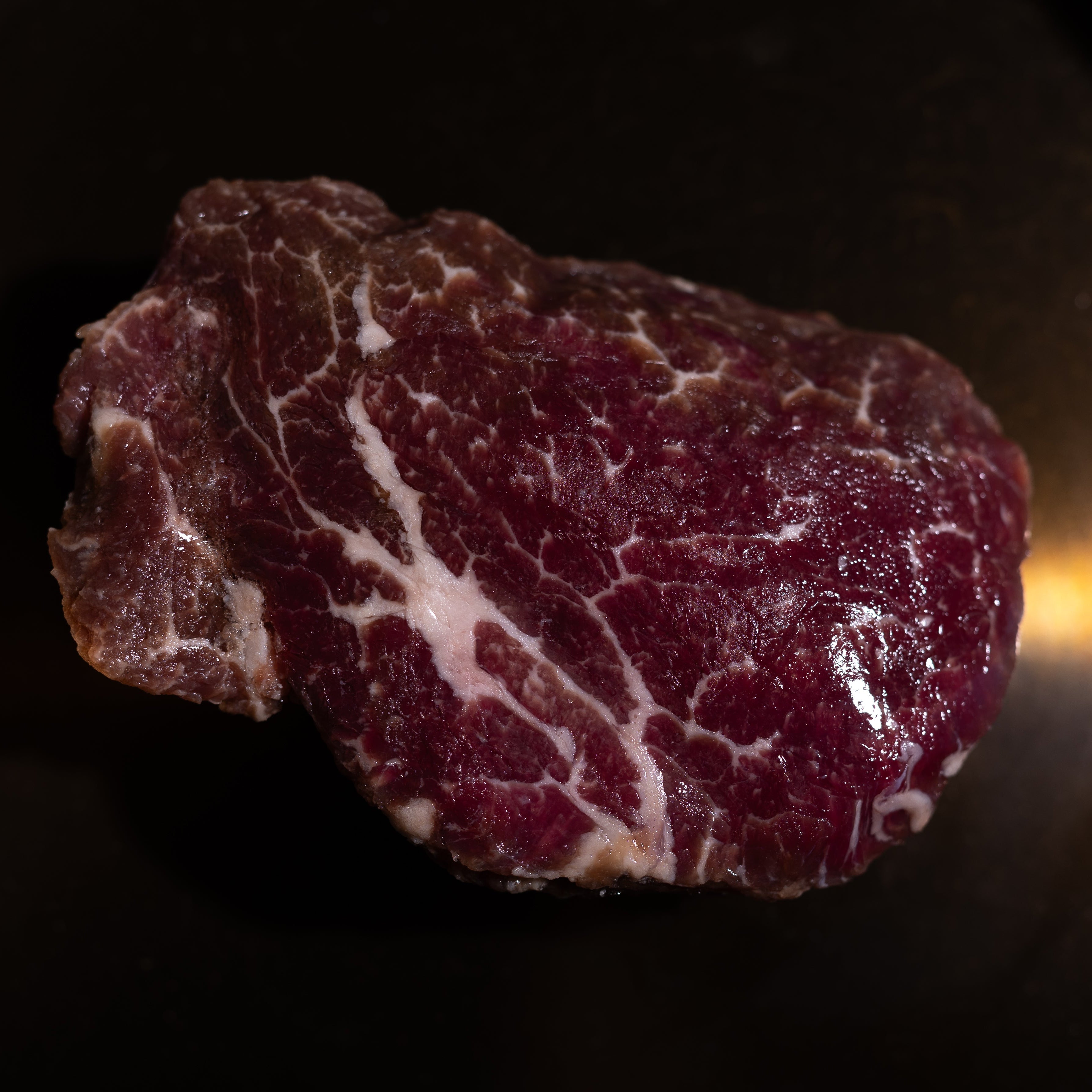 A raw marbled beef steak or cut of meat placed on a dark pan or plate with warm light reflecting off the surface.