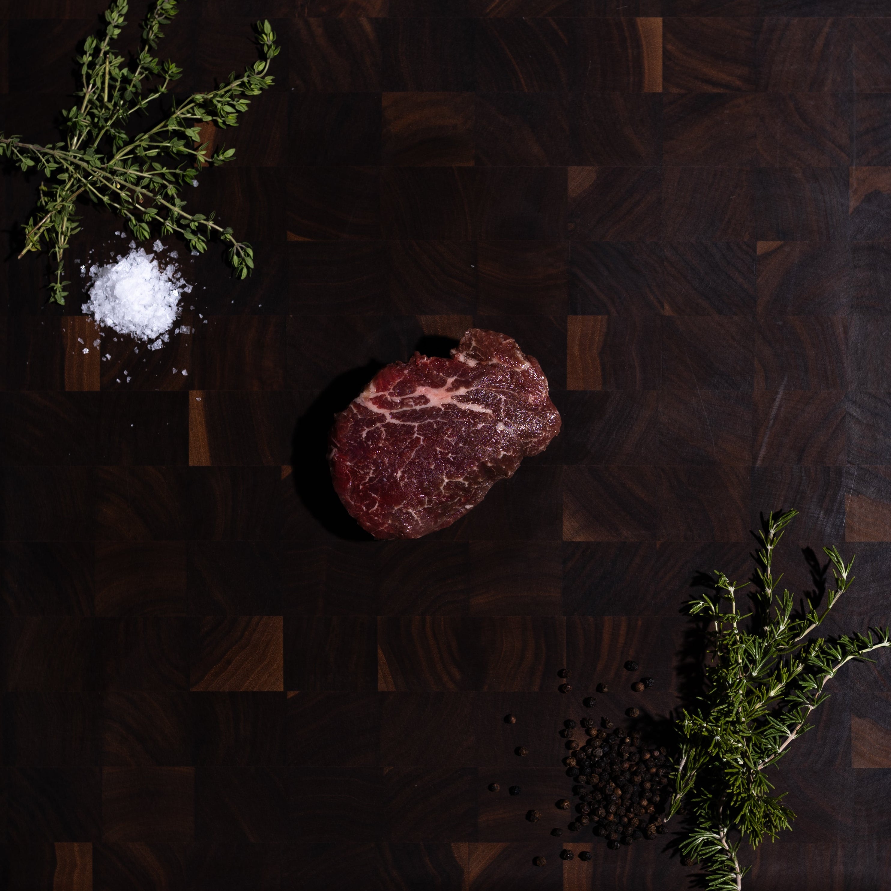 A raw cut of beef steak placed on a dark wood cutting board, styled with coarse salt, herbs, and scattered peppercorns on a dark green surface.