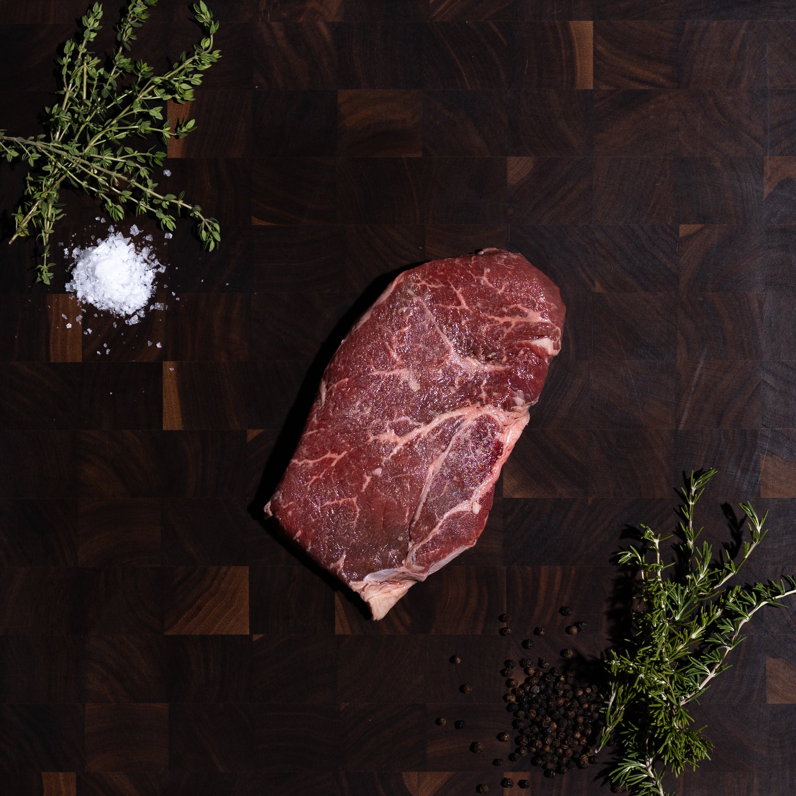 A raw marbled beef steak rests on a dark wooden cutting board, styled with a pinch of coarse salt, green herb sprigs, and black peppercorns on a dark olive-green surface.