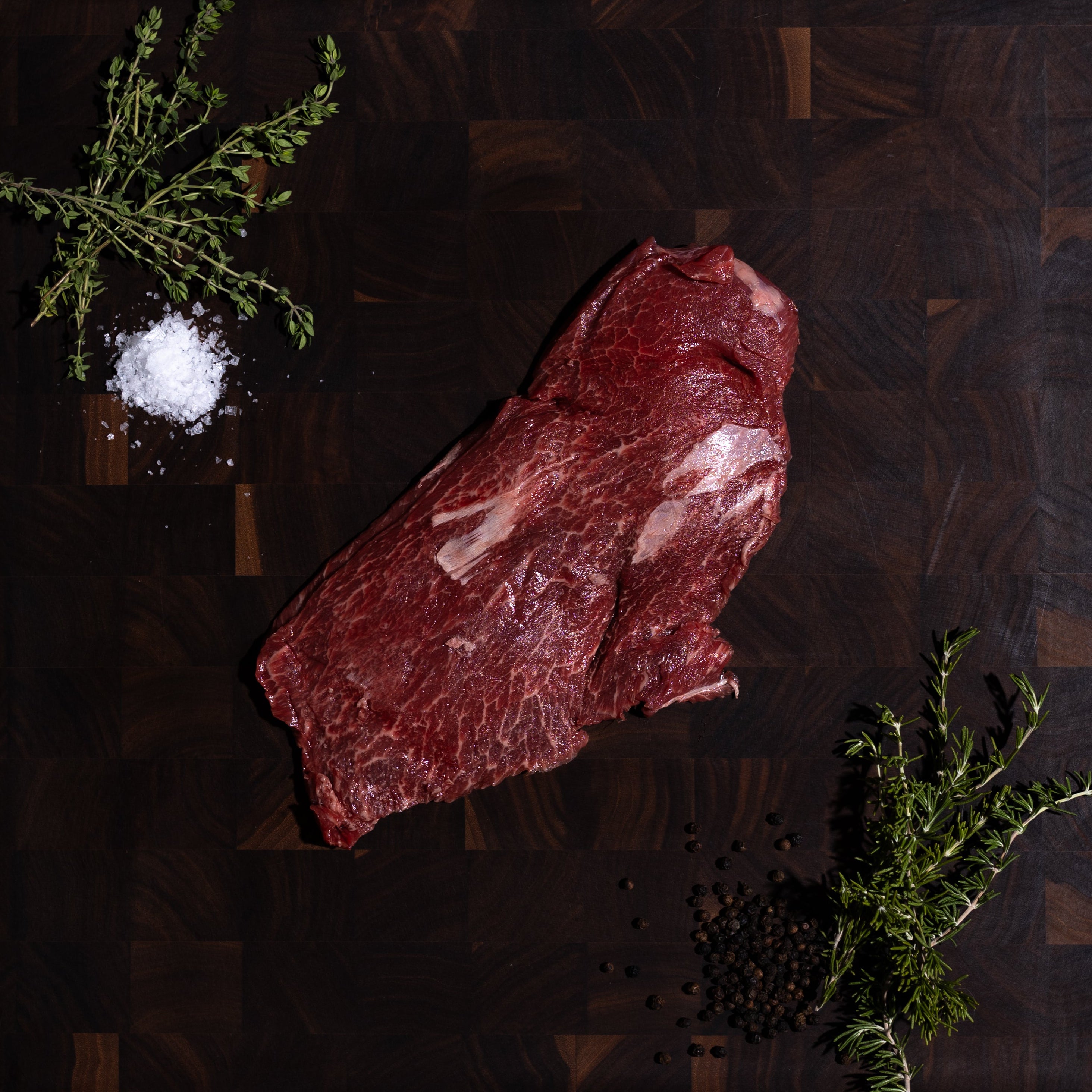 A raw cut of red beef steak placed on a dark wooden cutting board, styled with a sprig of rosemary, coarse sea salt, and black pepper on a dark green surface.