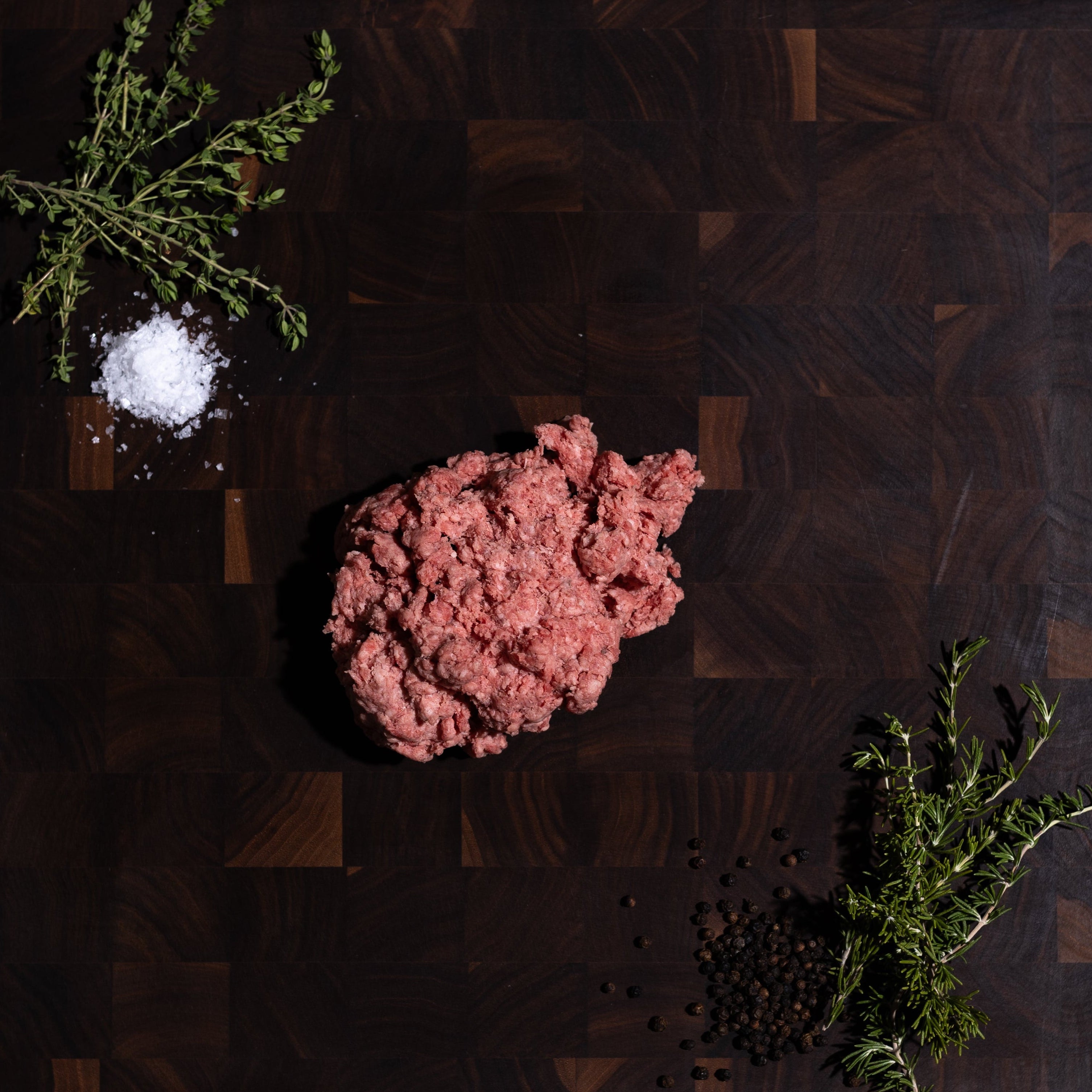 Raw ground meat placed on a dark wooden cutting board, with a small pile of white salt, green herb sprigs, and black peppercorns arranged around it on a dark green surface.