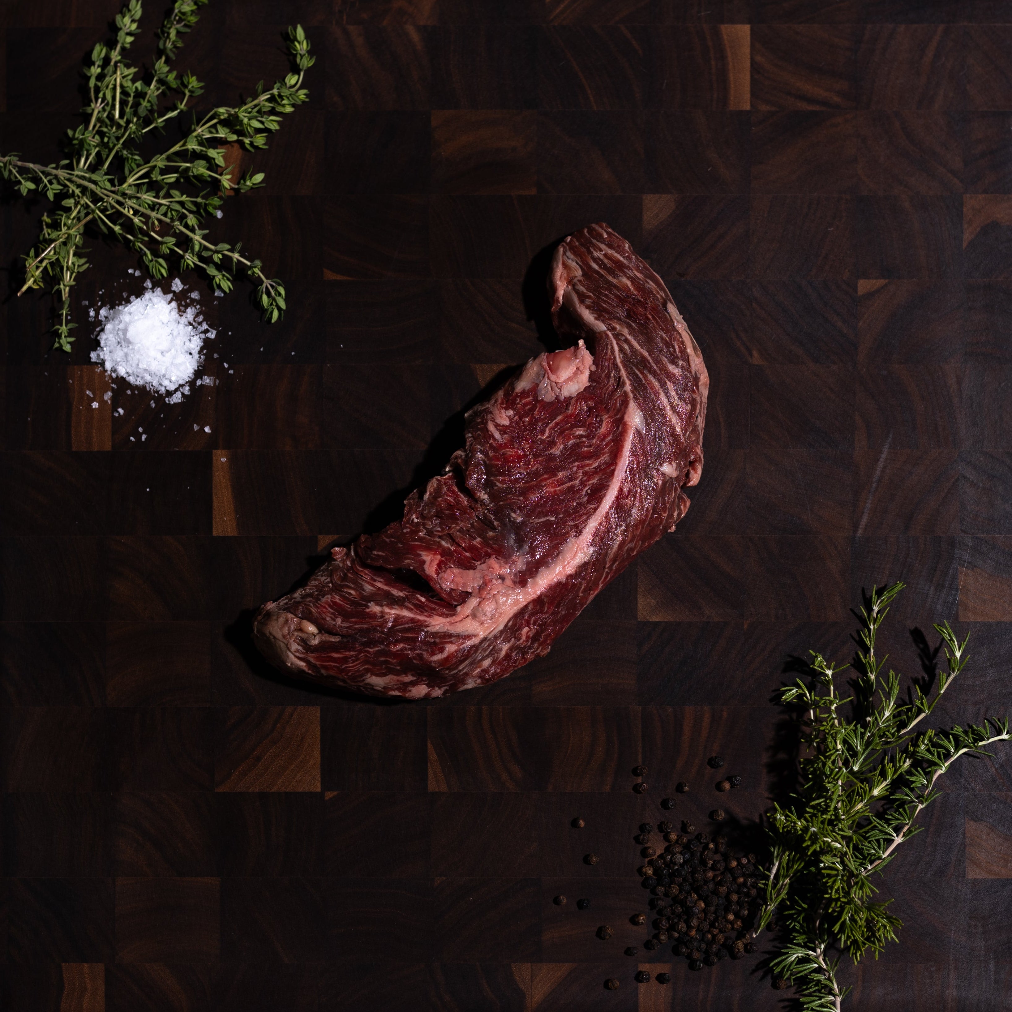 A raw cut of beef steak placed on a dark wooden cutting board, styled with a small pile of coarse salt, a sprig of herb, and scattered peppercorns on a dark green surface.