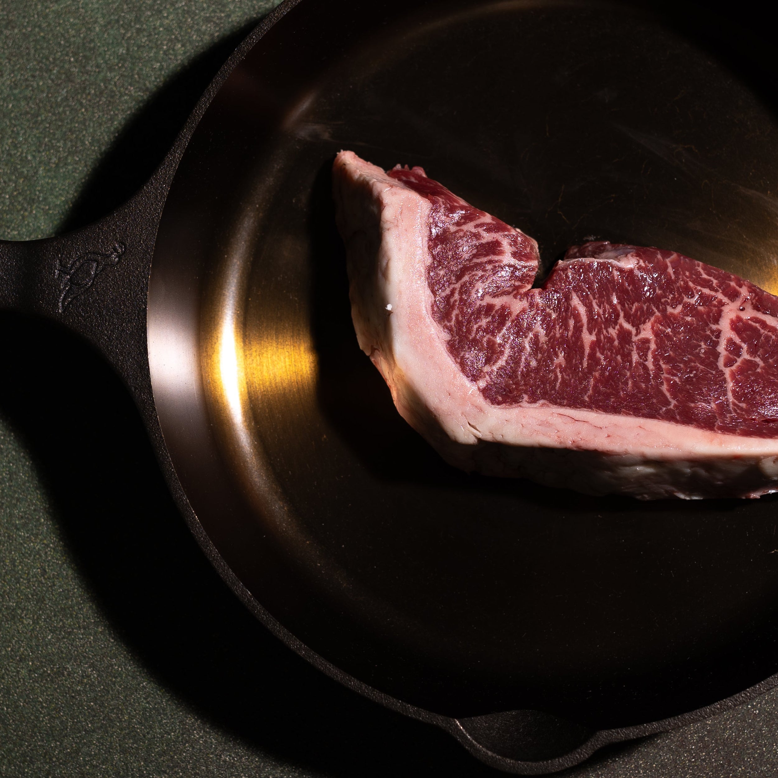 A raw marbled beef cut sits inside a black cast iron skillet on a dark textured background.