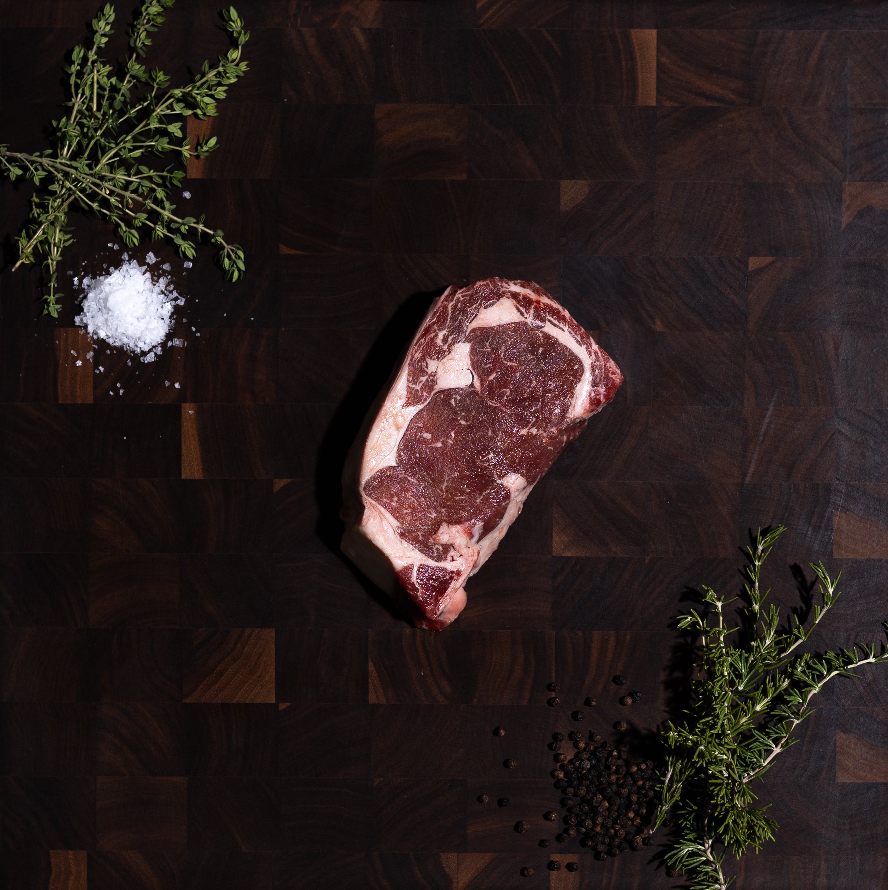 A raw marbled steak rests on a dark wooden cutting board, styled with a sprig of rosemary, coarse salt, and black peppercorns on a dark green textured background.