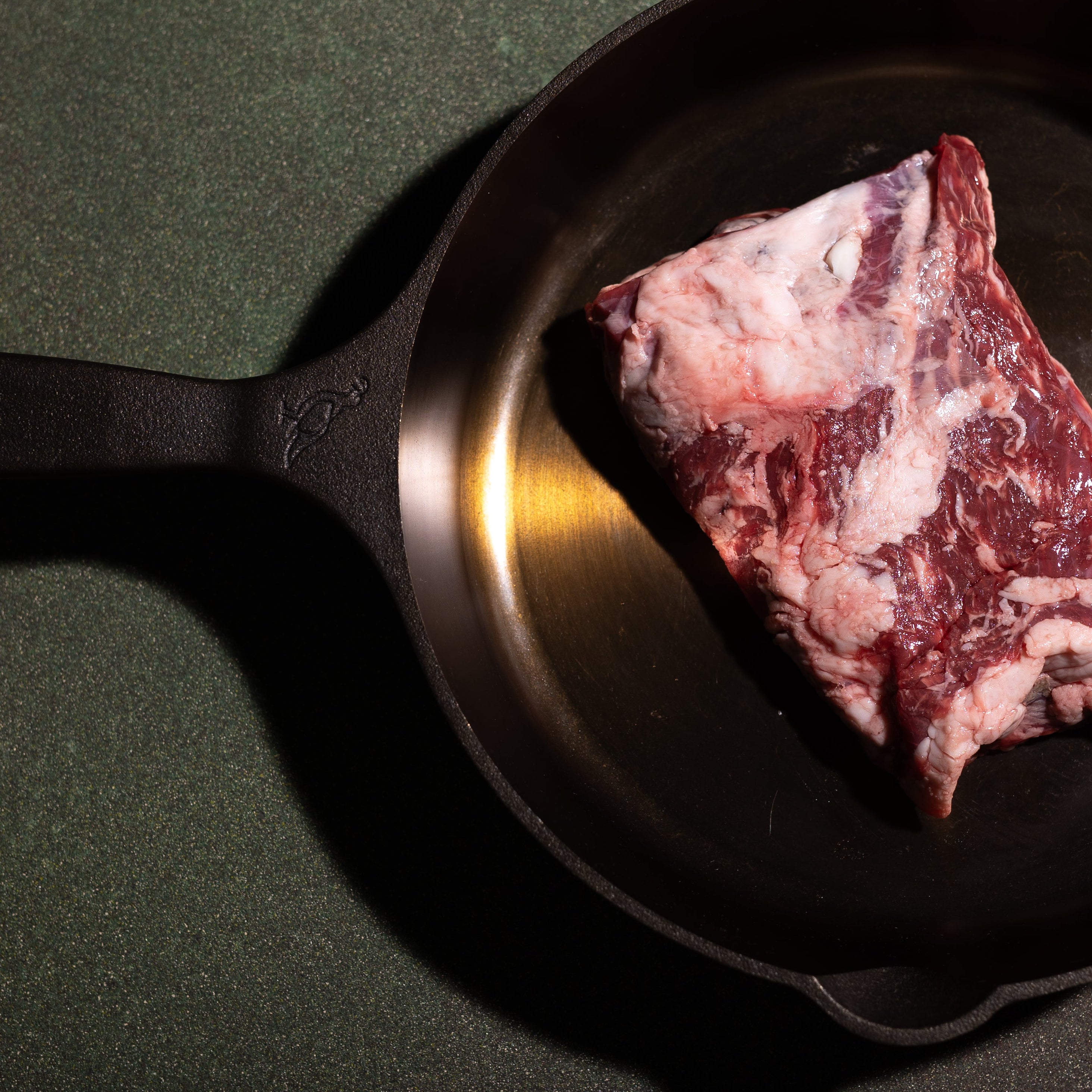 A raw cut of marbled meat sits inside a black cast iron skillet on a dark green textured surface.