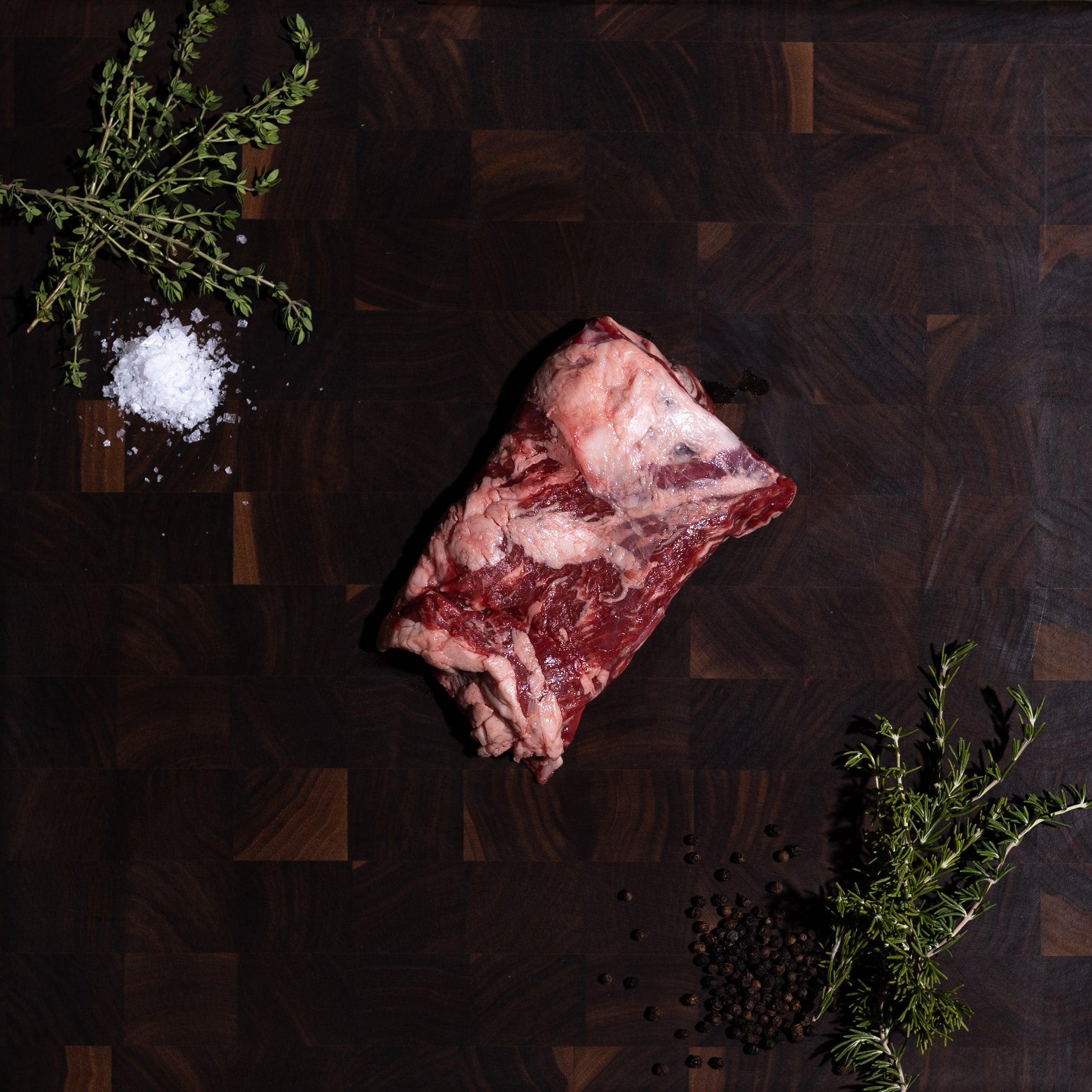 A raw cut of marbled meat placed on a dark wooden cutting board, styled with a small pile of coarse salt, herbs, and peppercorns on a dark green surface.