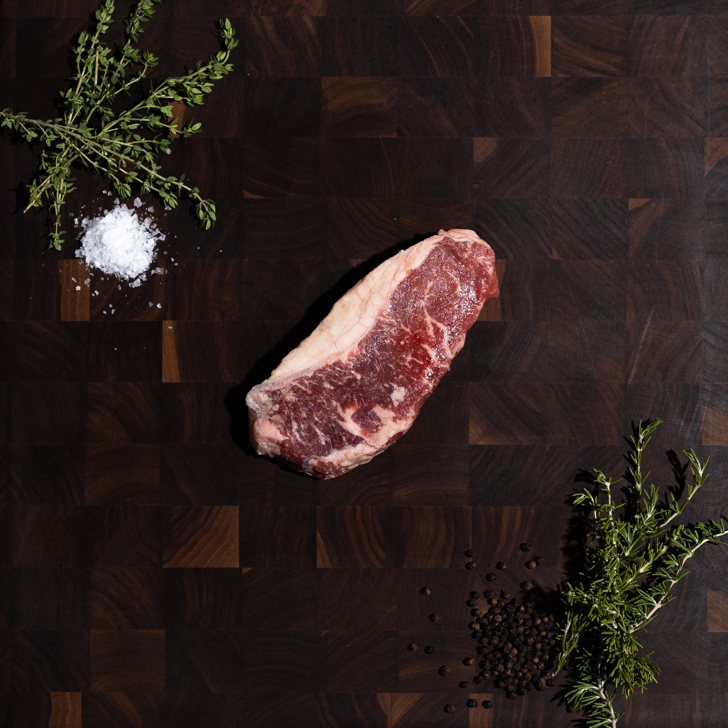 A raw marbled beef steak rests on a dark wood cutting board, styled with a sprig of rosemary, coarse salt, and black peppercorns on a dark green textured background.