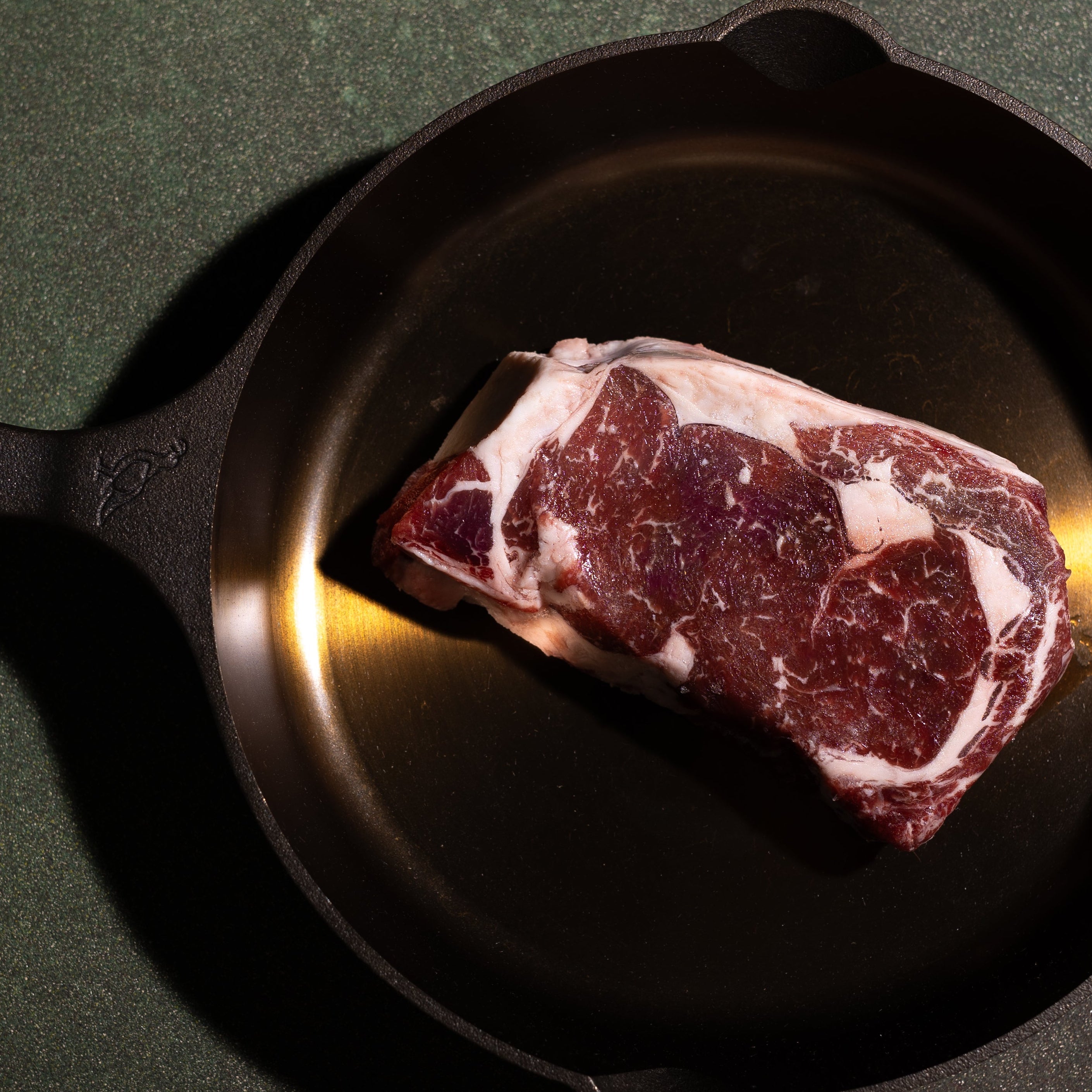 A raw cut of marbled steak placed inside a black cast iron skillet on a dark green textured surface.