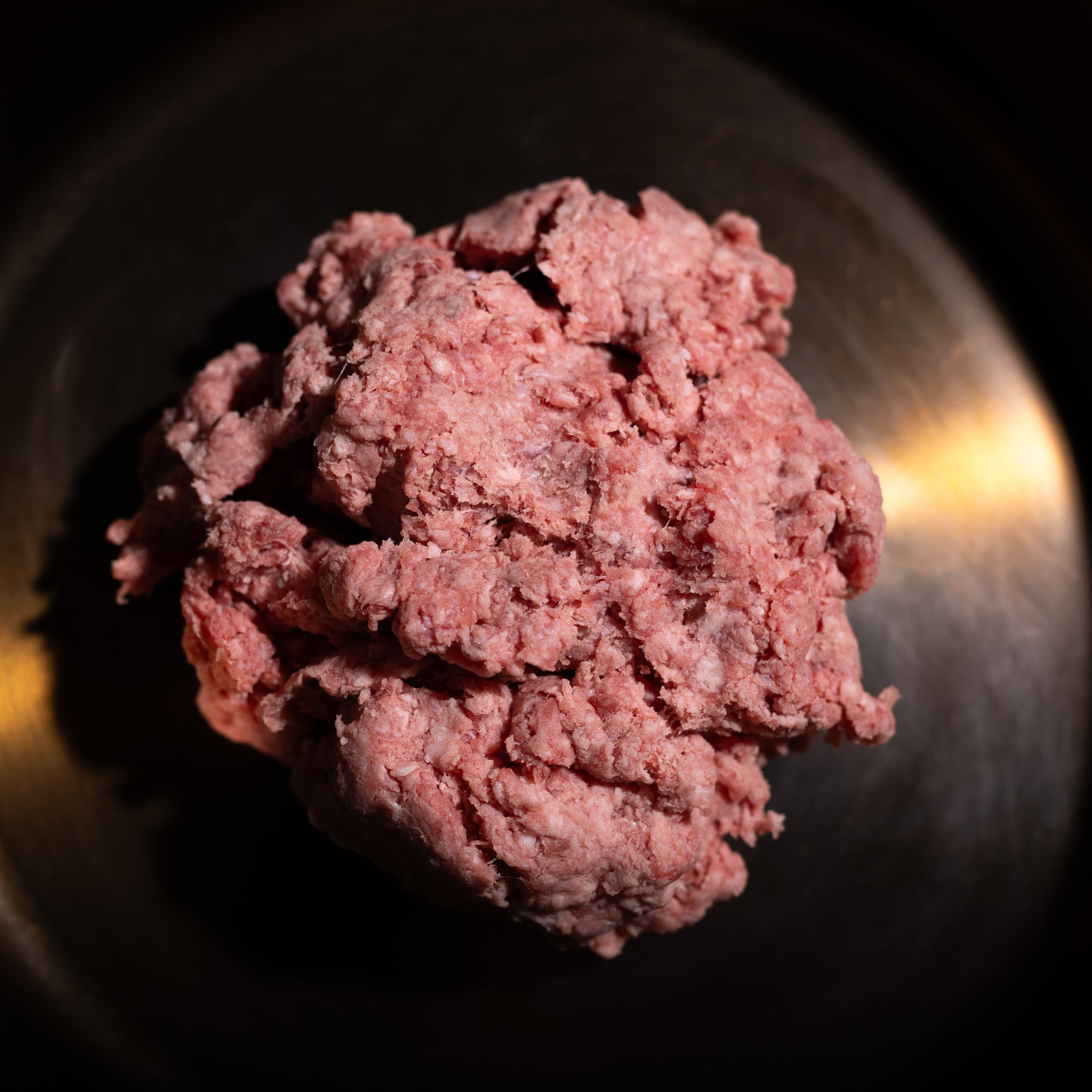 Raw ground meat, likely beef or pork, placed in the center of a dark cast iron skillet on a dark background.