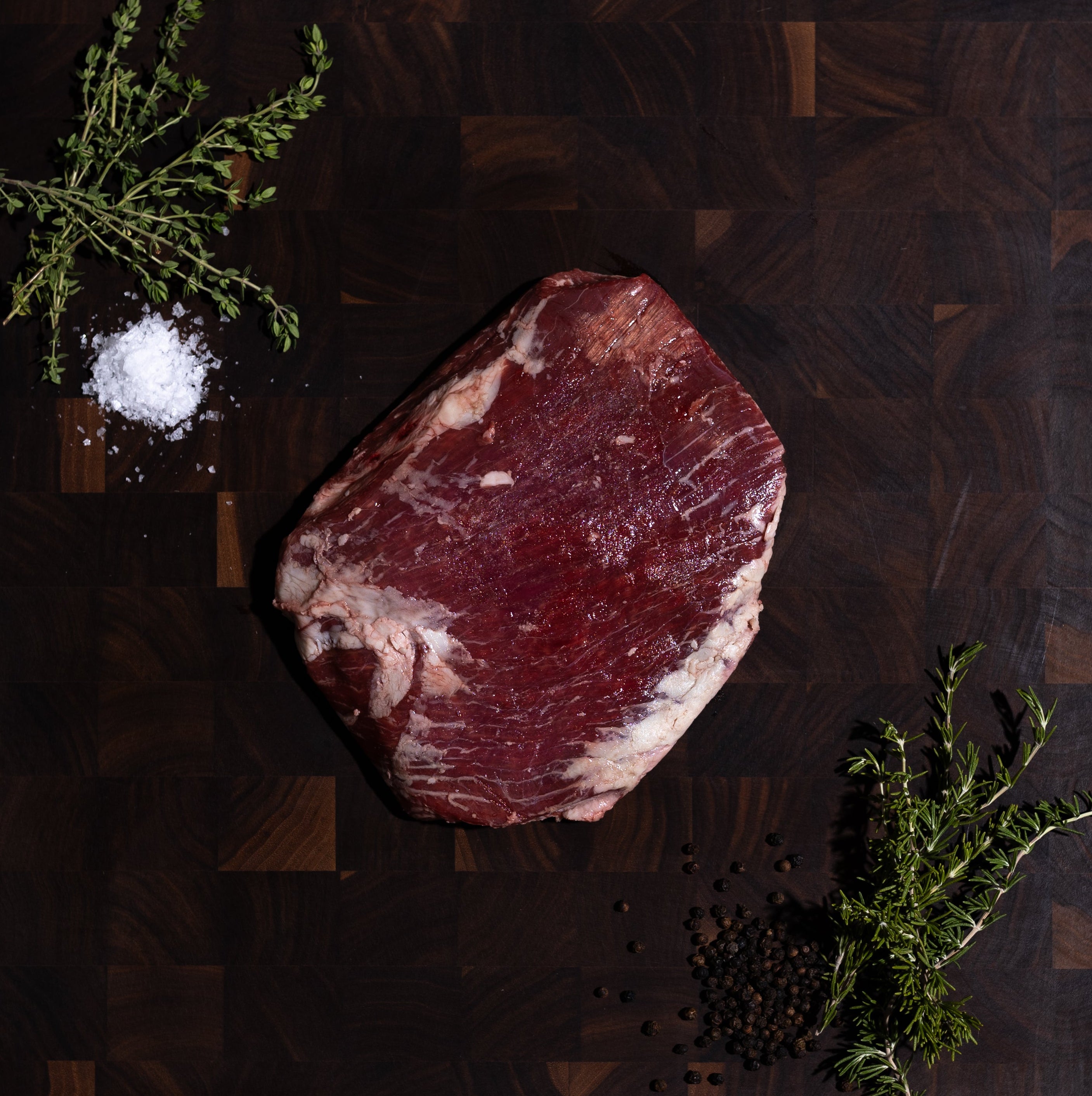 A raw cut of red meat with fat marbling sits on a dark wooden cutting board, styled with a pinch of coarse salt, black peppercorns, and green herb sprigs on a dark green surface.