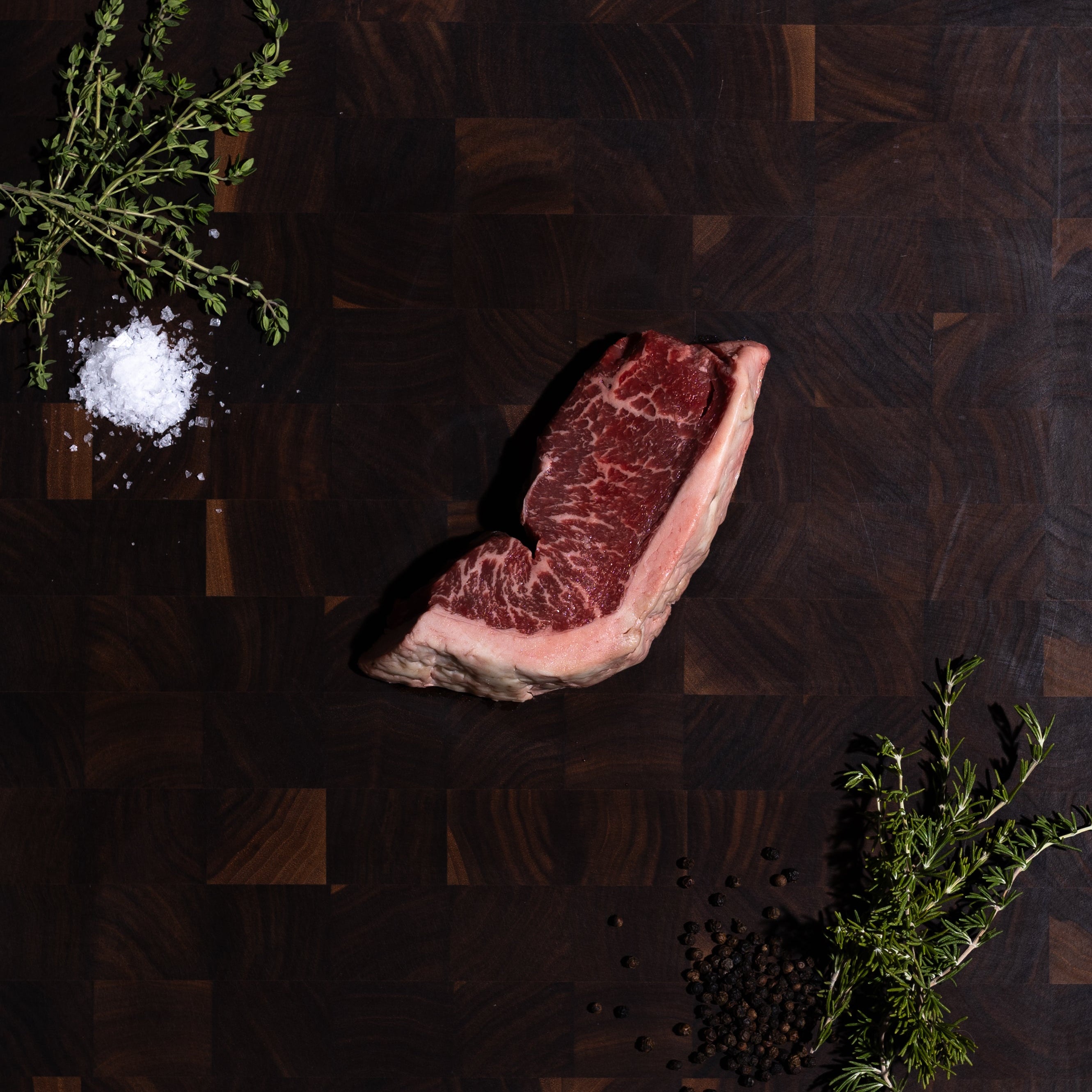 A raw beef steak with marbling is arranged on a dark wooden cutting board, styled with a sprig of herbs, coarse salt, and peppercorns on a dark green background.