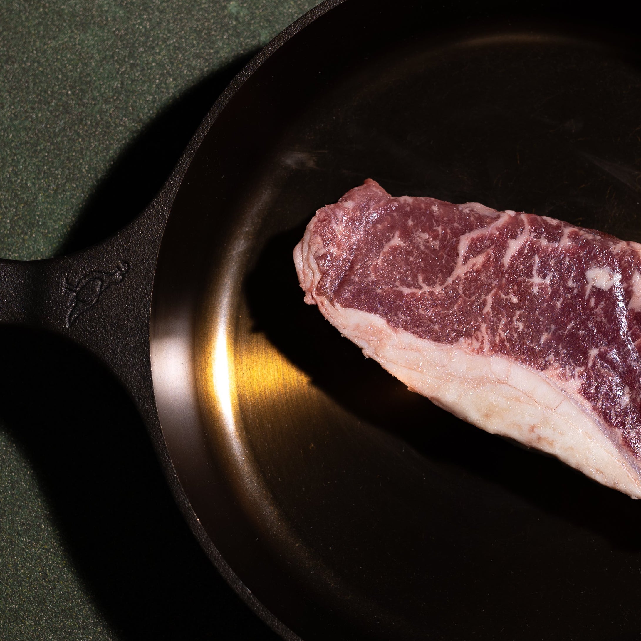 A raw cut of marbled beef rests inside a black cast iron skillet on a dark green textured surface.
